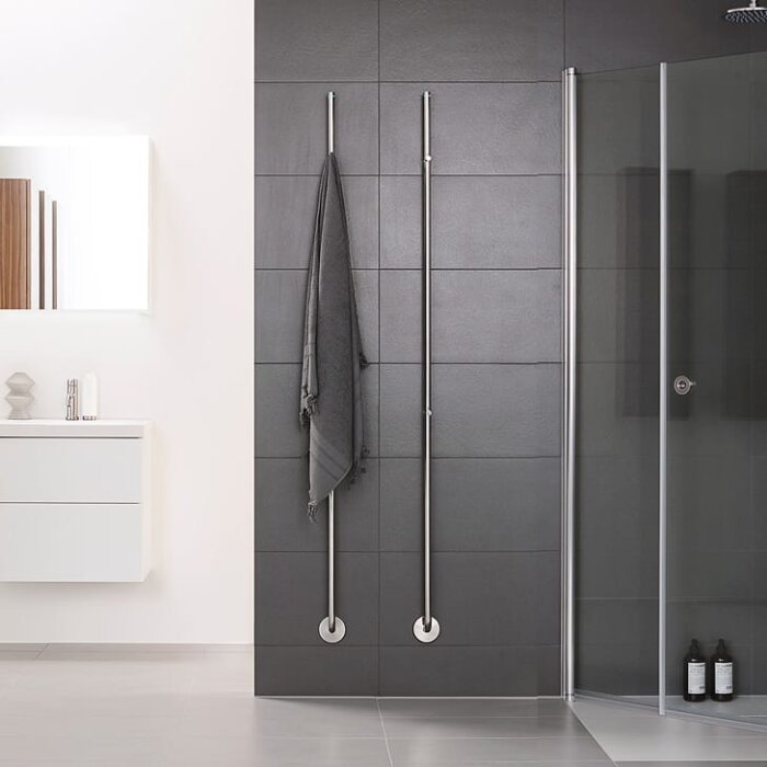 Två lodräta handdukstorkar monterade på en mörk kaklad badrumsvägg, en med en hängande grå handduk.