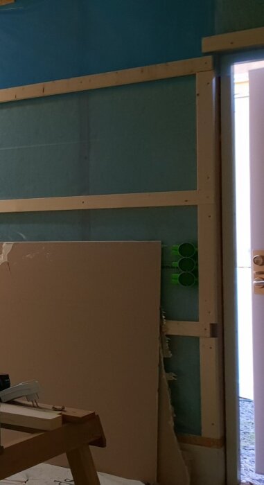 Vågräta lister fastsatta runt ett dörrhål på en trävägg med gipsskiva lutad mot väggen och arbetsbänk i förgrunden.