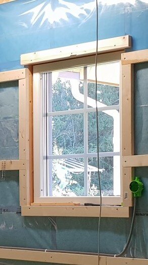 Träram fäst runt fönsteröppning, med glespanel och synlig isolering. Skog syns genom fönstret.