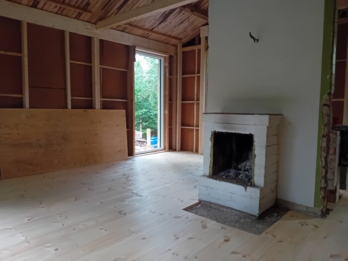 Öppen spis i ljust trärum under renovation, delvis rivet golv nära spisen, stora fönster öppnar mot utsidan med grönt landskap.