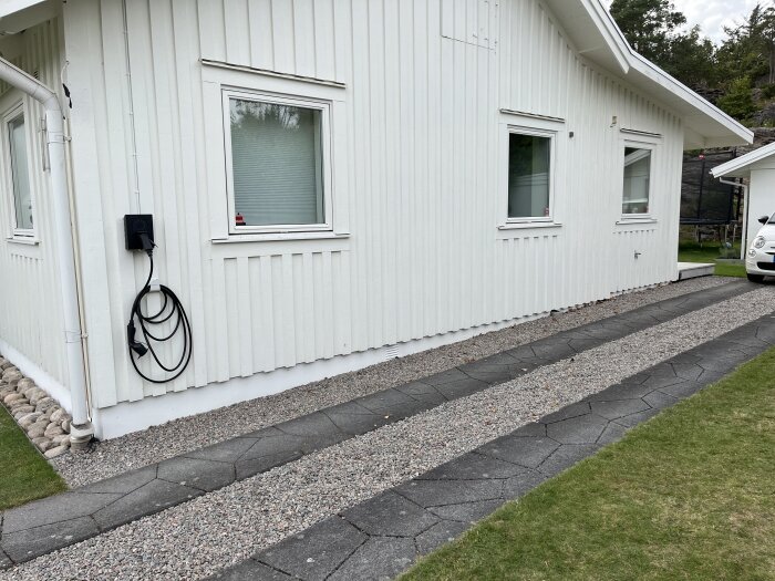 Vit träfasad av ett hus med grusad gång och stengång intill, elektrisk laddstation synlig på väggen.