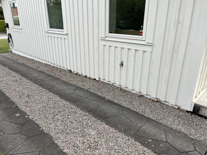 Vit fasad av hus med grus och betongplattor längs grunden; diskussion om dräneringsbehov vid eventuell ny uppfart planeras.