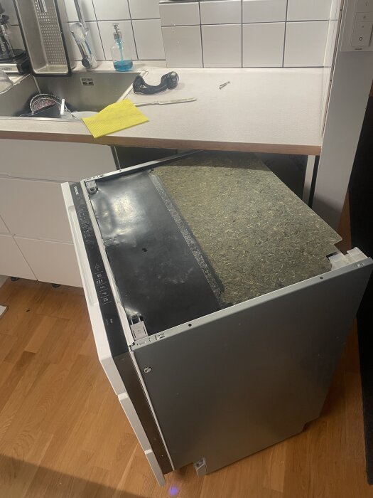 Inbyggd diskmaskin utdragen ur köksbänk för reparation, med verktyg och rengöringsmedel på bänkskivan.