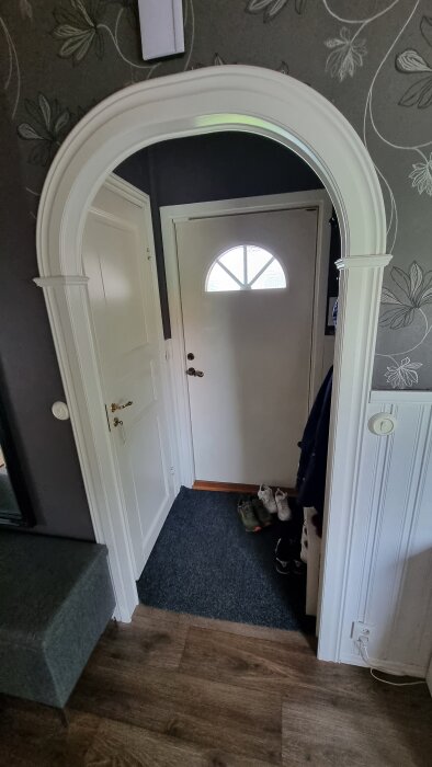 Vit dörr med runt fönster, mörk matta, skor och blommigt tapetmönster i hall.