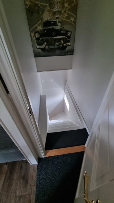 Vit trappa sedd från ovan med tavla föreställande en bil på väggen ovanför. En dörrkarm och ett ganska mörkt rum visas också.