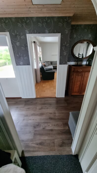 Utsikt från hall med trägolv och spegel mot vardagsrum med soffa, delvis skymd bild.