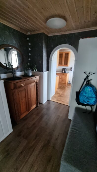 Entré med mörkt trägolv, en byrå med spegel, väggbeklädnad och valv in till kök. Resväska hänger på en krok.