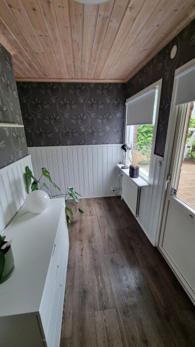 Trångt rum med mörkt trägolv, vit panelvägg och blommig tapet. Fönsterbräda med lampa och växt, vit byrå med krukväxt.