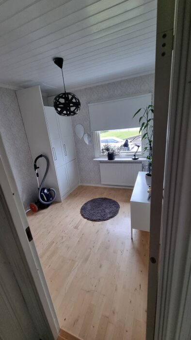 Ljust rum med trägolv, vit garderob, dammsugare, runda speglar, och fönster med utsikt över trädgård. Växt och lampa på bord under fönstret.