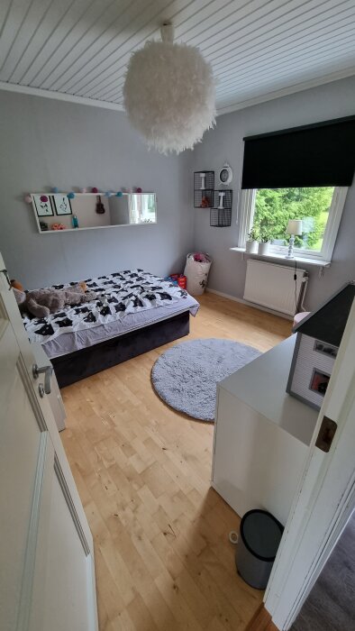 Ett sovrum med grå väggar, säng med mönstrat överkast, rund matta, fjäderlampa, och hylla med prydnadssaker. Grönt utanför fönstret.