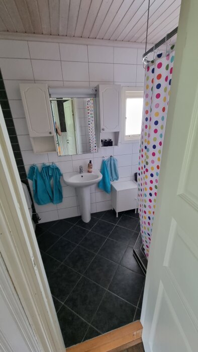 Badrum med vitt kakel, svart golv, färgglad duschdraperi och handfat. En del av bilden är delvis skymd av ett finger.