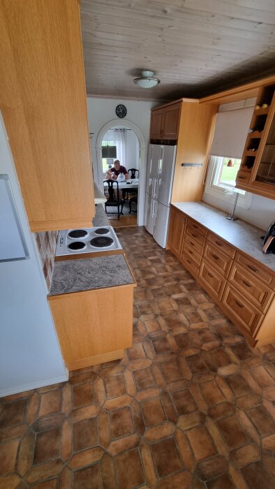 Kök med trägolv och träskåp, spis och kylskåp synliga; spegel reflekterar en person som sitter vid ett bord i bakgrunden.