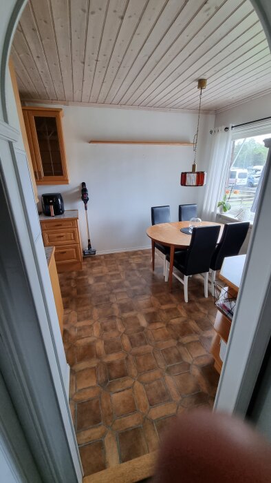 Kök med träpaneltak, brun golvmatta, ett runt matbord med fyra stolar och ett skymmande finger i nedre kanten.