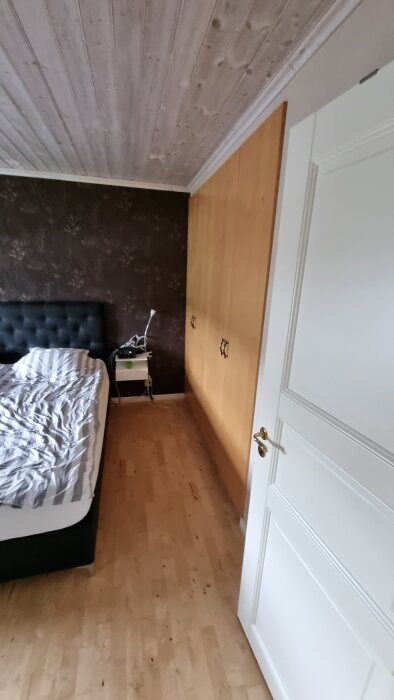Sovrum med mörka väggar, trägolv och tak, en säng närmast kameran samt en garderob längs väggen.