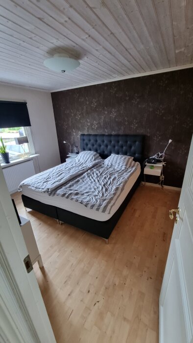 Sovrum med dubbelsäng, brunt blommönstrat fondvägg, ljust trägolv och vitt tak med lampa.