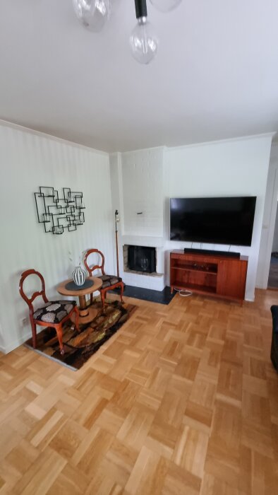 Vardagsrum med parkettgolv, öppen spis, TV, två stolar och ett runt bord, delvis skymt av ett finger.