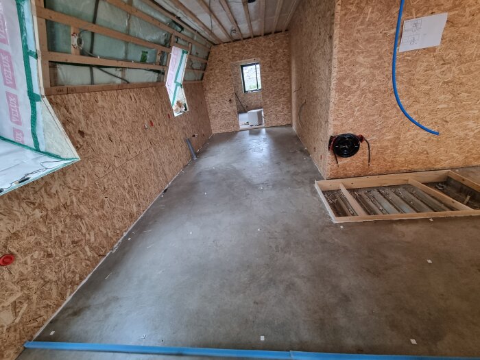 Nyspacklat golv i ett rum med träfiberskivor på väggarna och synliga träbjälkar i taket under byggprocessen.