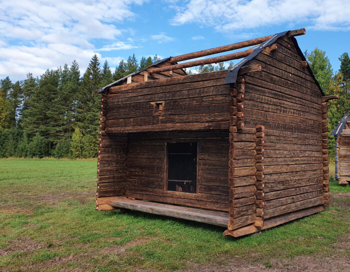 Nedplockat härbre från 1877 i stockar, på en gräsplan med skog i bakgrunden.
