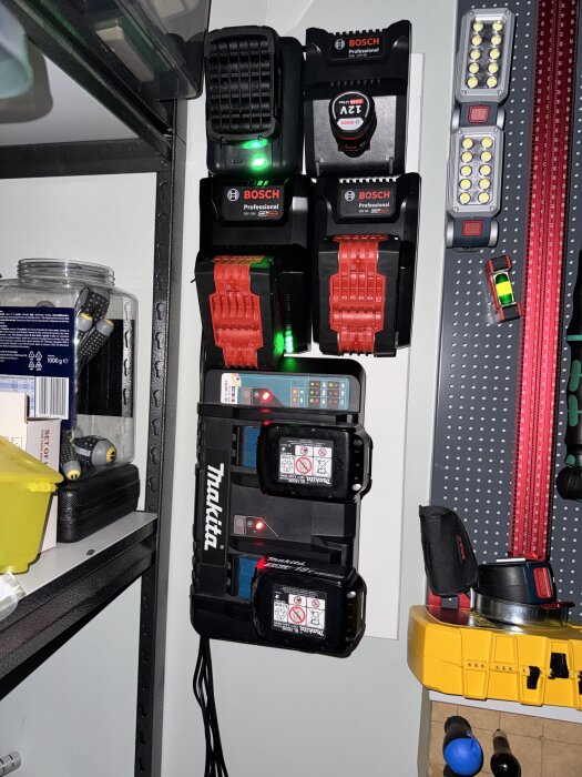 Väggmonterade batteriladdare från Bosch och Makita med flera batterier, omgivna av verktyg och utrustning i en verkstad.