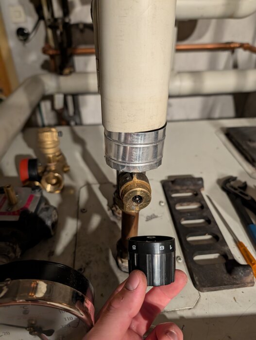 Svart vred i en hand, avlossnat från ett rör med synlig mässingsnippel. Verktyg och rördetaljer syns i bakgrunden.