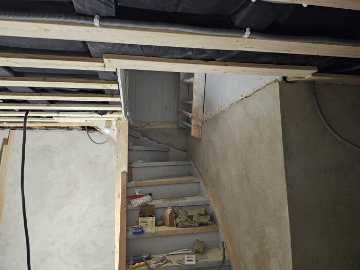 Renovering av källare med obehandlade trägolv på trappan, synliga kablar och flexislang i taket samt gipsskivor under montage.
