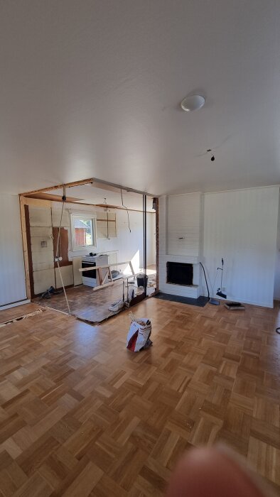 Renovering pågår i vardagsrum och kök, med nedplockade väggar och blottat tak. En vit eldstad syns till höger och en ICA-påse ligger på parkettgolvet.