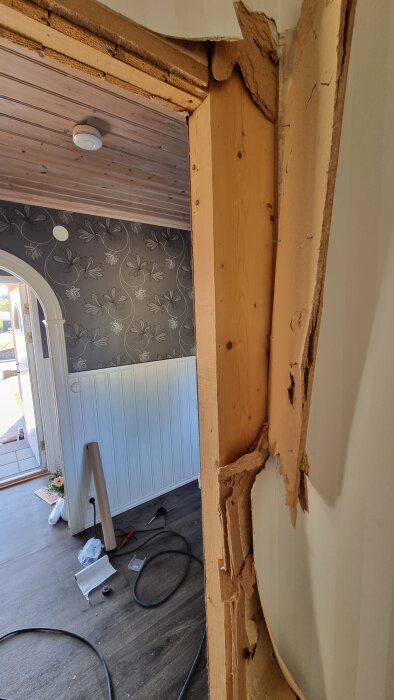Demonterad dörrkarm med synliga träreglar och skadad väggbeklädnad i ett rum under renovering, sladdar och verktyg ligger på golvet.