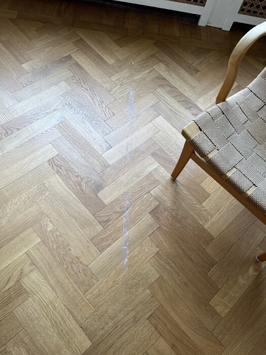 Parkettgolv med maskeringstejprester i ett solbelyst rum, försök att ta bort, stol synlig till höger.