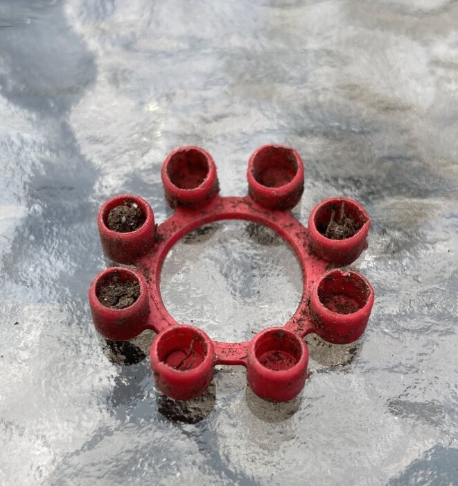 Röd plastdel, rund med åtta rörformade öppningar fyllda med jord. Hittad vid trädgårdsgrävning.