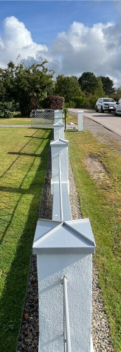 Nymålat, vitt staket med stålkant och hattar på stolparna, omgivet av gräsmatta och grus, under en klarblå himmel med moln.