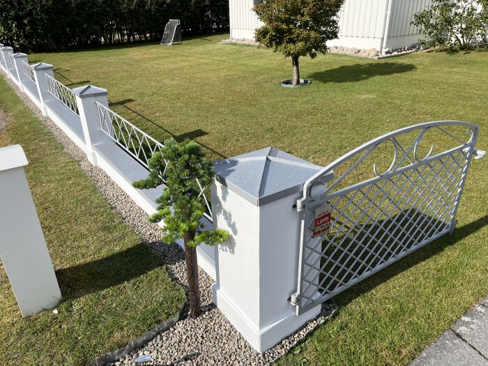 Nyrenoverat vitt staket med metalldetaljer och konstgjord tall, stenbelagt fundament och belysning på grindstolpar, mot en grön gräsmatta.