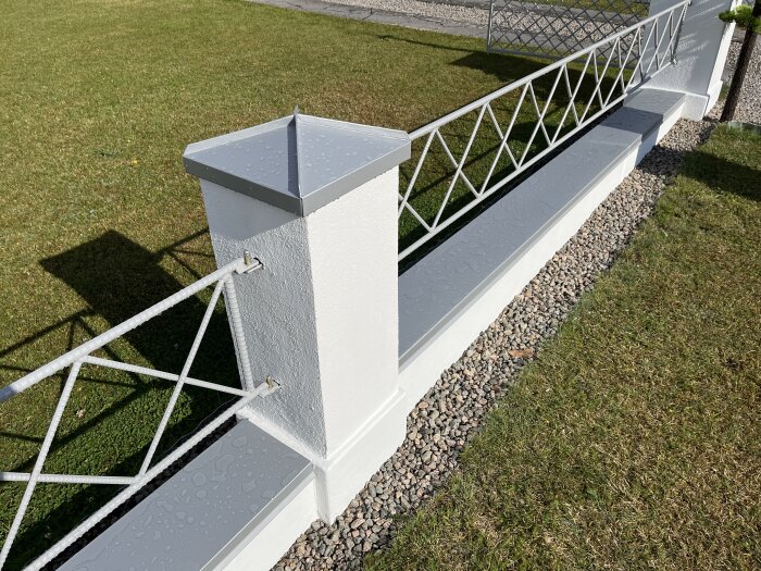 Vitmålat staket med metallstänger och betongmur, kantat med grus. Solbelyst med skuggor på gräsmatta.