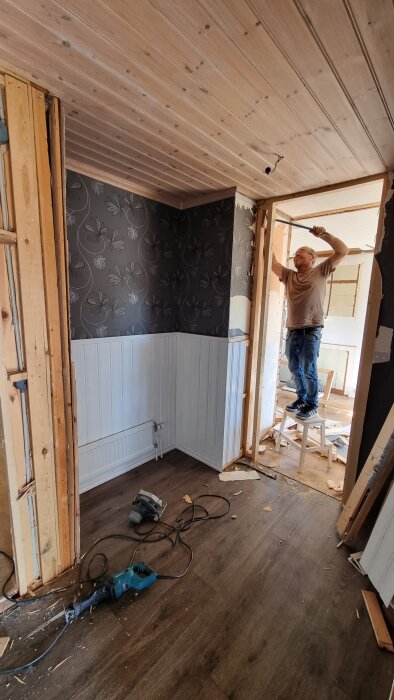 Renovering pågår i rum med blomtapet; man står på pall och arbetar på ramar mellan nedtagna väggar. Verktyg på golvet.