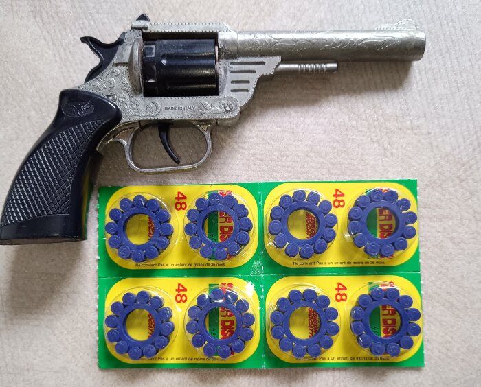 Leksakspistol i metall med svarta handtag, bredvid ligger sex förpackningar med smällkappladdningar på en beige yta.