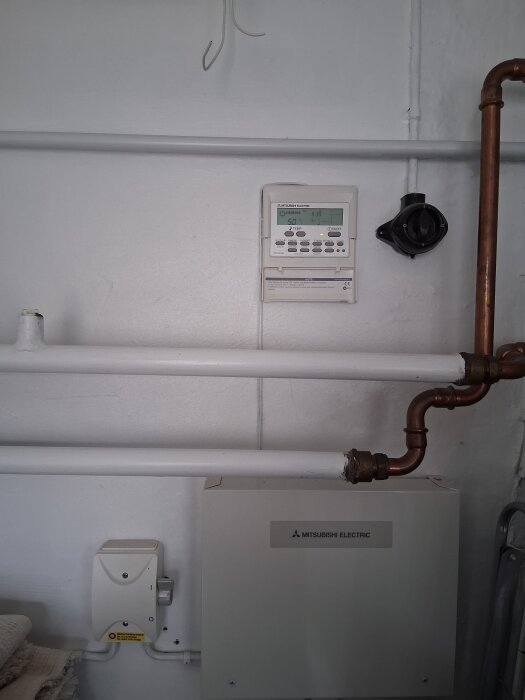 Luftvärmepump från Mitsubishi Electric installerad på en vit vägg med synliga rör och kontrollpanel, potentiellt i pannrum.