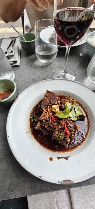 Grissida serverad i ett djupt fat med mörk sås, garnerad med koriander och chili. Ett glas rött vin står bredvid på bordet.