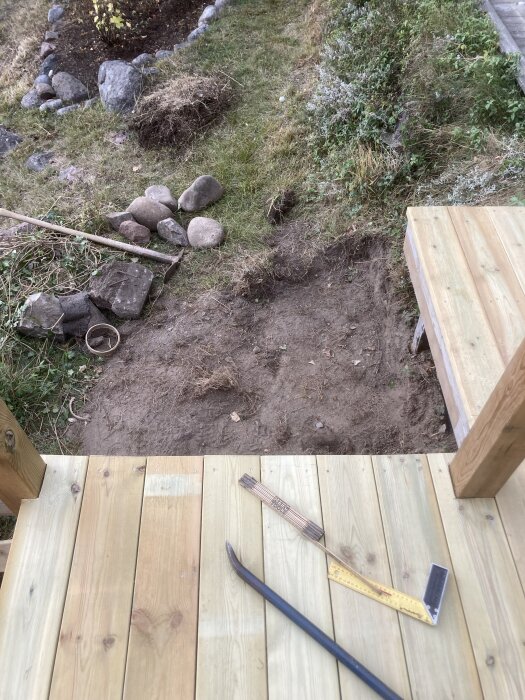 Altan med måttband, kofot och snickarvinkel. Jordigt område och stenar syns framför altanen, med gräs och buskar på sidorna.