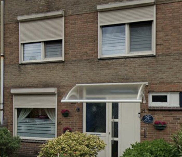 Fasad av tegelhus med fönsterjalusier och entrétak, omgiven av växter och hängblommor.