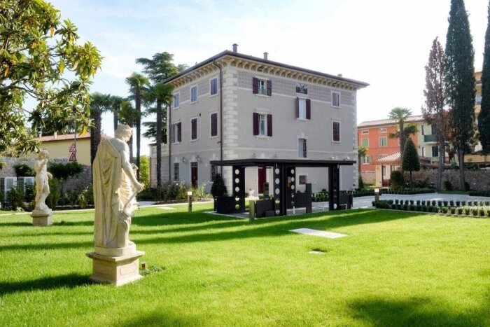 Stort stenhushus i liberty stil med fönsterluckor, omgiven av en välskött trädgård med statyer och grönt gräs.