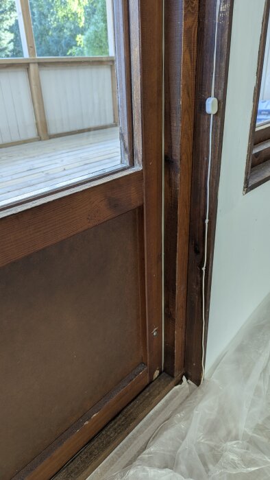 Närbild av brun träaltandörr med synliga åldringsmärken, del av inre karm, och utsikt mot veranda med trädäck.