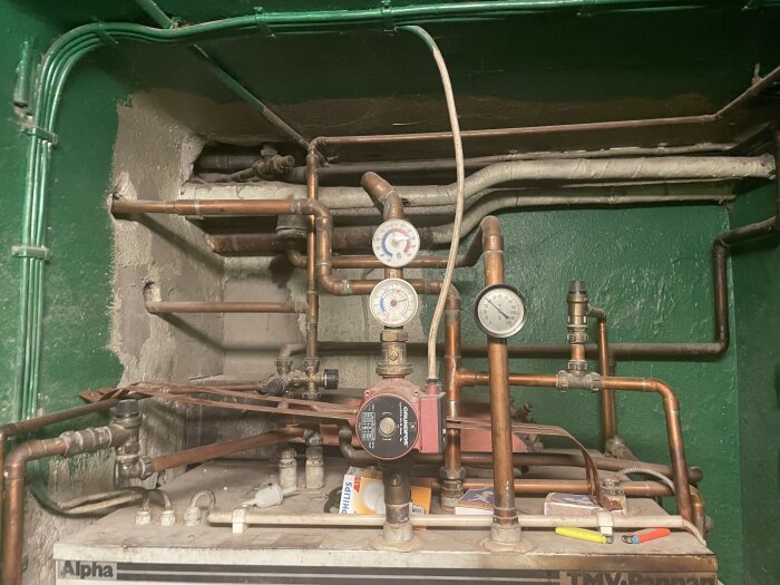 Äldre kombinationspanna med rör och ventiler, tryckmätare visar 1 och 2 bar. På märkningen syns "Alpha" och "TMV". Verktyg och tändsticksaskar på pannans ovansida.
