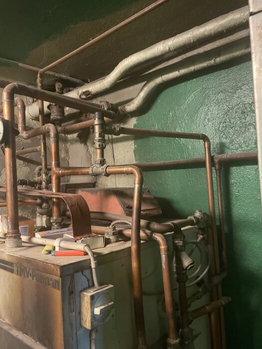 Äldre TMV kombinationspanna med flera kopparrör och ventiler installerad mot en grön vägg. Verktyg och föremål ligger ovanpå pannan.