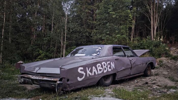 Gammal bil med skador och texten "SKABBEN" målad på sidan, stående i en grön skogsmiljö.