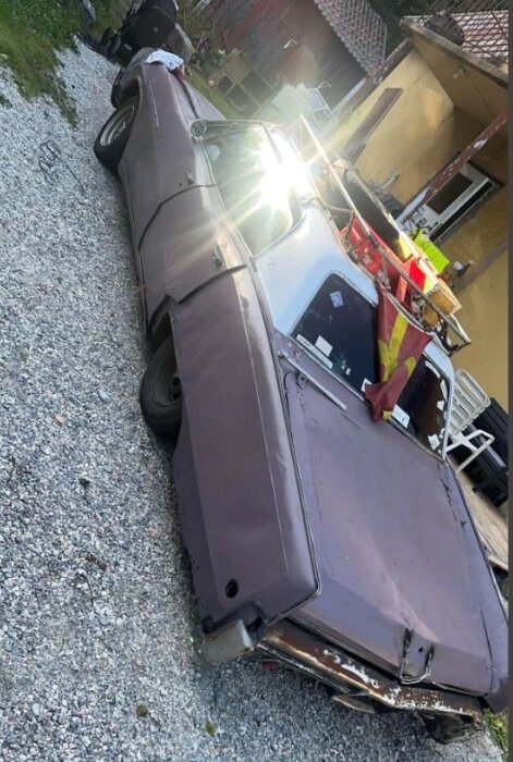 Rostig bil med bucklor står parkerad på en grusuppfart, omgiven av verktyg och reservdelar, med solstrålar reflekterande på bilens yta.