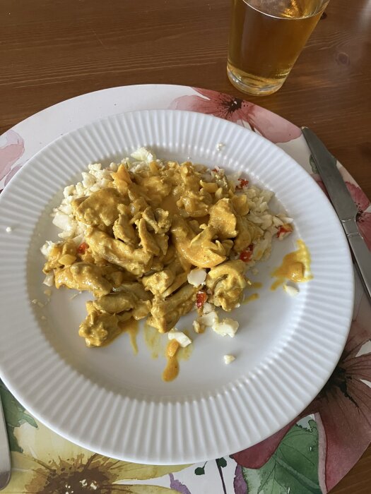 Kycklinggryta med curry, chili och créme fraîches serverad med blomkålsris med paprika på en vit tallrik, med ett glas dryck i bakgrunden.