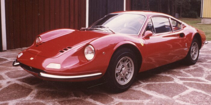 En röd sportbil parkerad på stenlagd uppfart framför en träbyggnad.