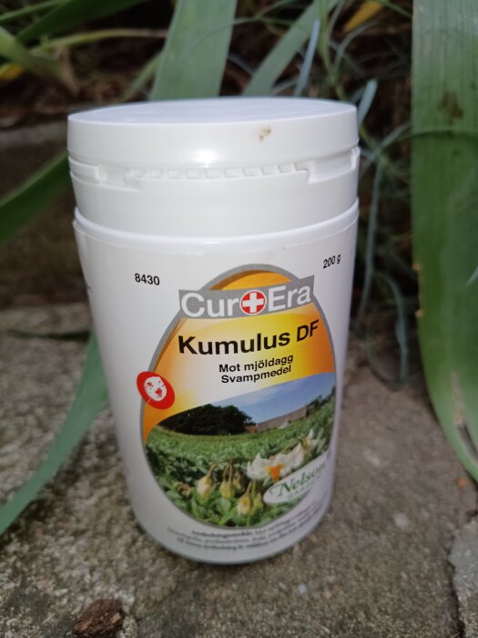 Behållare med svampmedlet Kumulus DF från Nelson Garden, avsett för bekämpning av mjöldagg, står på en stenig yta med gröna växter i bakgrunden.