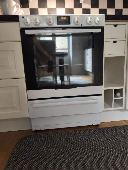 Vit spis med digital klocka och vred för inställningar, står i ett kök med svartvitt kakel.