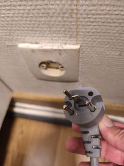 Grå elkontakt hålls framför ett smutsigt vägguttag på vägg med struktur.