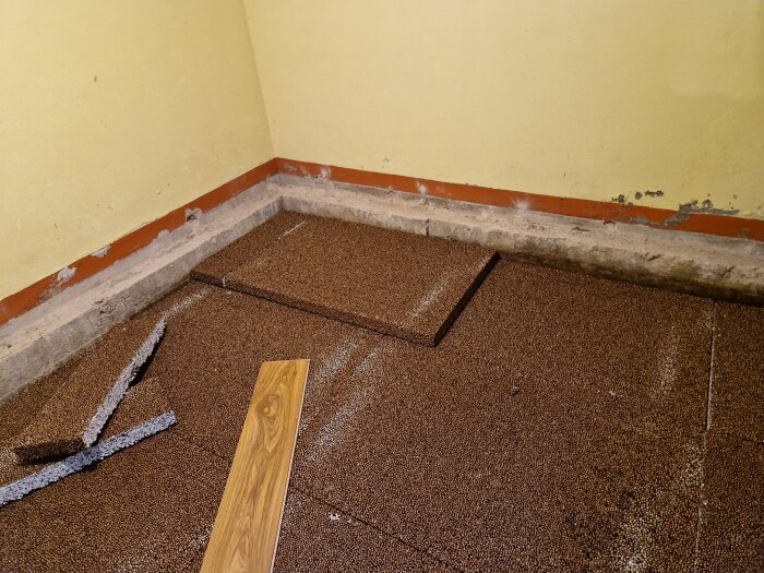 Golvläggning med bruna plattor och en ljus trälist vid en vägg med gul tapet och slitet hörn.